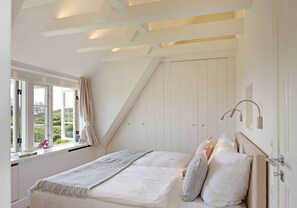 Ein Schlafzimmer mit schrägem Dach, einem großen Fenster mit Vorhängen, einem Bett mit Kissen und einer Nachttischlampe.