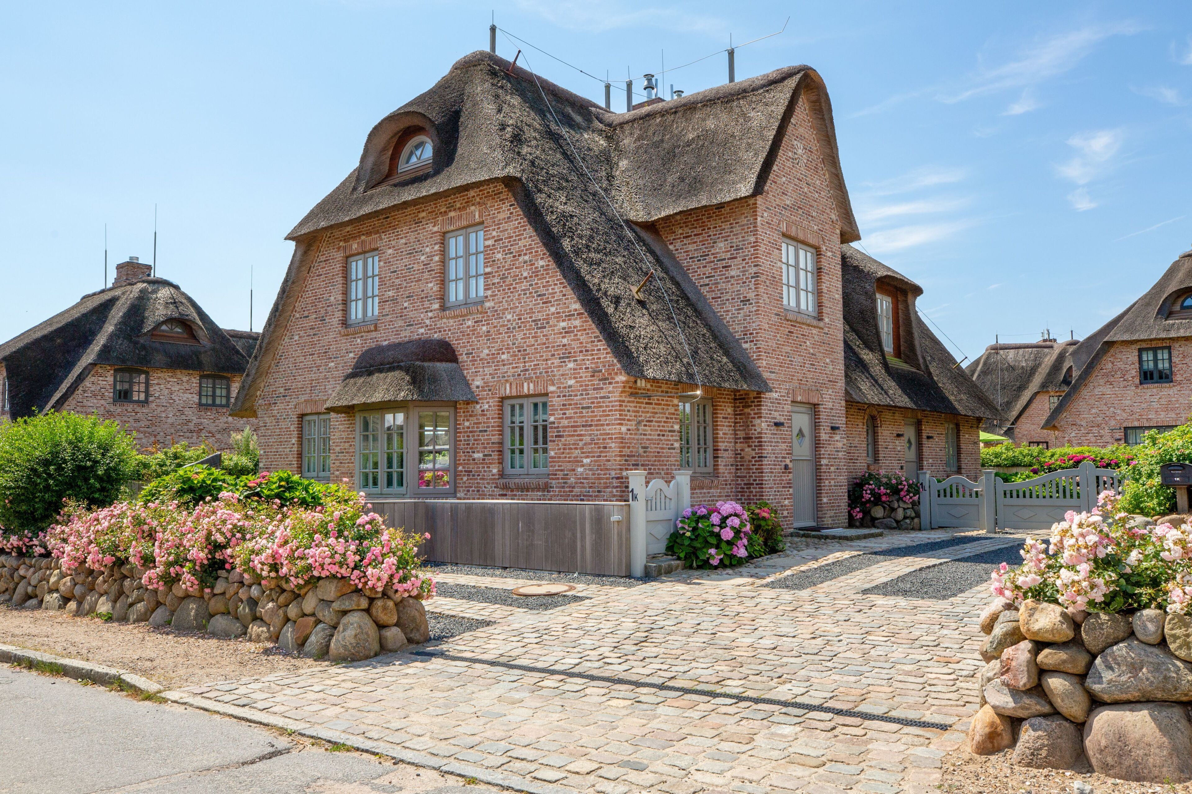A traditional brick house with a steeply pitched thatched roof, a stone wall with pink flowers, and a paved driveway.