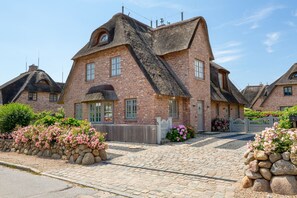 Une maison traditionnelle en briques, dotée d’un toit de chaume à forte pente, d’un mur de pierres orné de fleurs roses et d’une allée pavée.