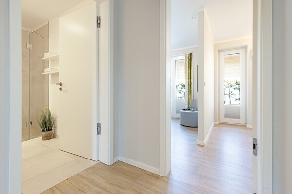 A modern hallway with a glass shower, a potted plant, and a view of a living area with a large window and a chair.