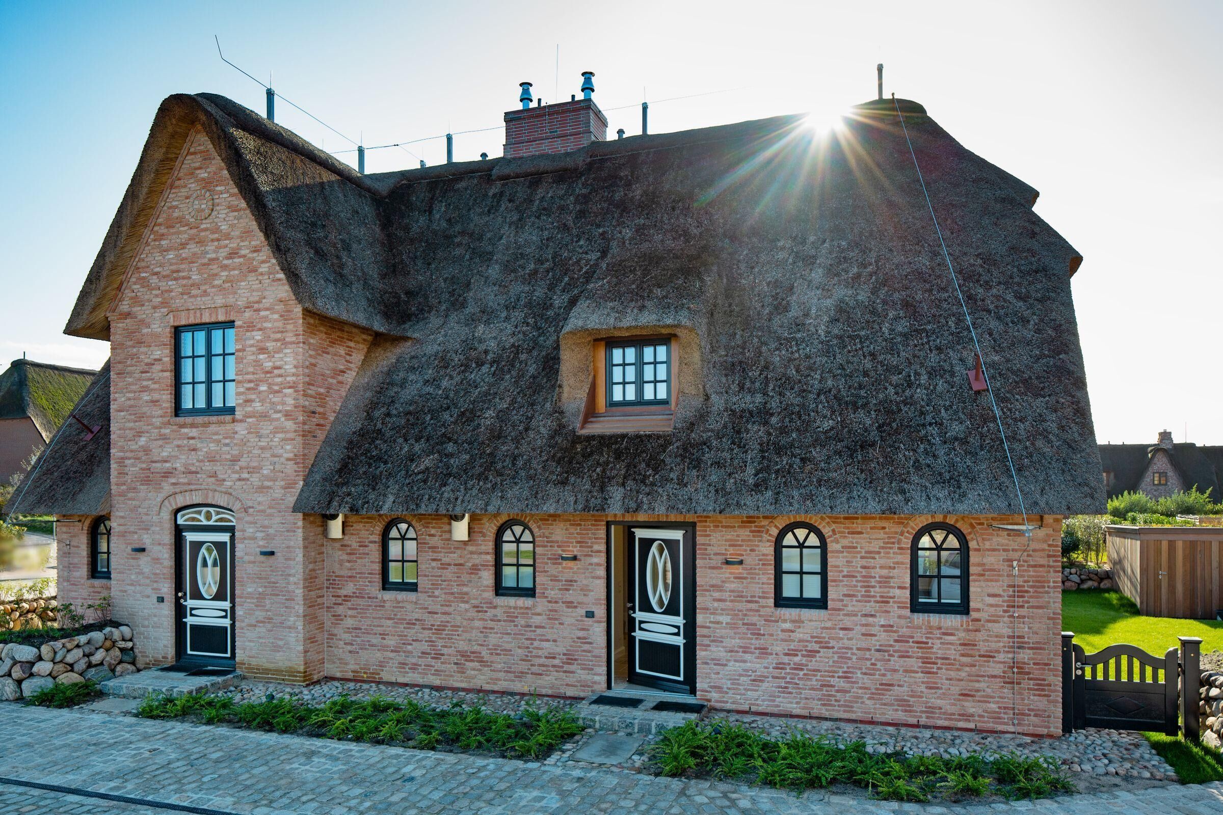 A traditional brick house with a steeply pitched thatched roof, multiple windows, and a well-maintained garden.