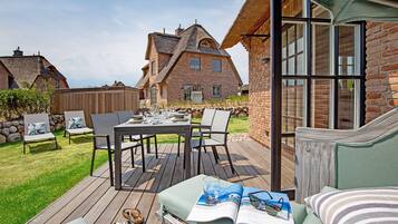An outdoor patio with a wooden deck, a dining table set for four, and a green sofa with a magazine and sunglasses on it.