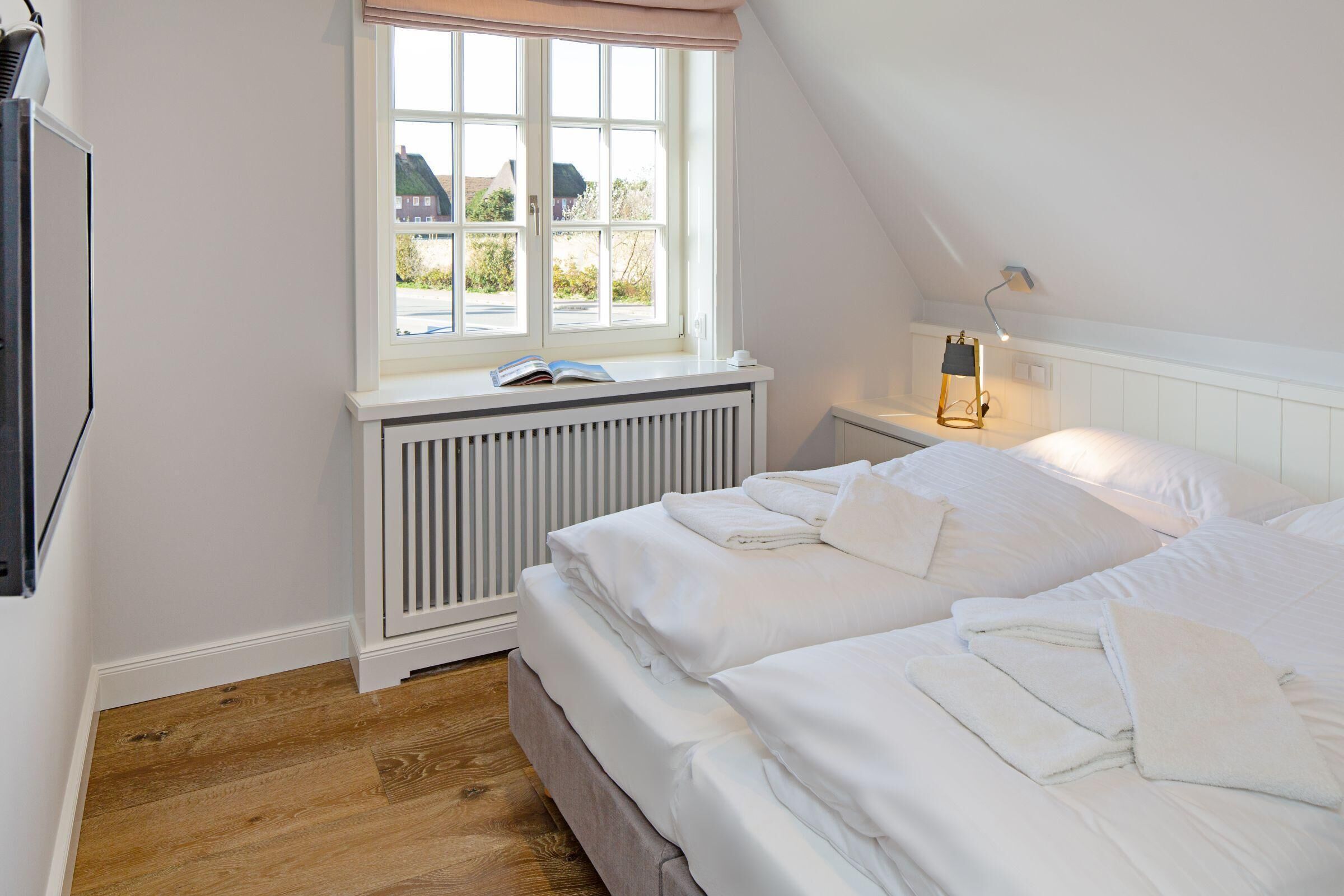 Une chambre avec une grande fenêtre, un radiateur, une table de chevet avec une lampe et des lits soigneusement faits.