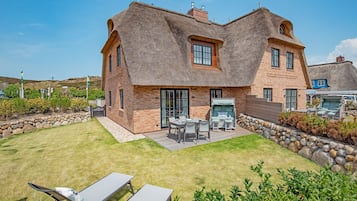 Een huis met rieten dak, bakstenen muren en een stenen fundering. Er is een terras met tuinmeubilair en een gazon.