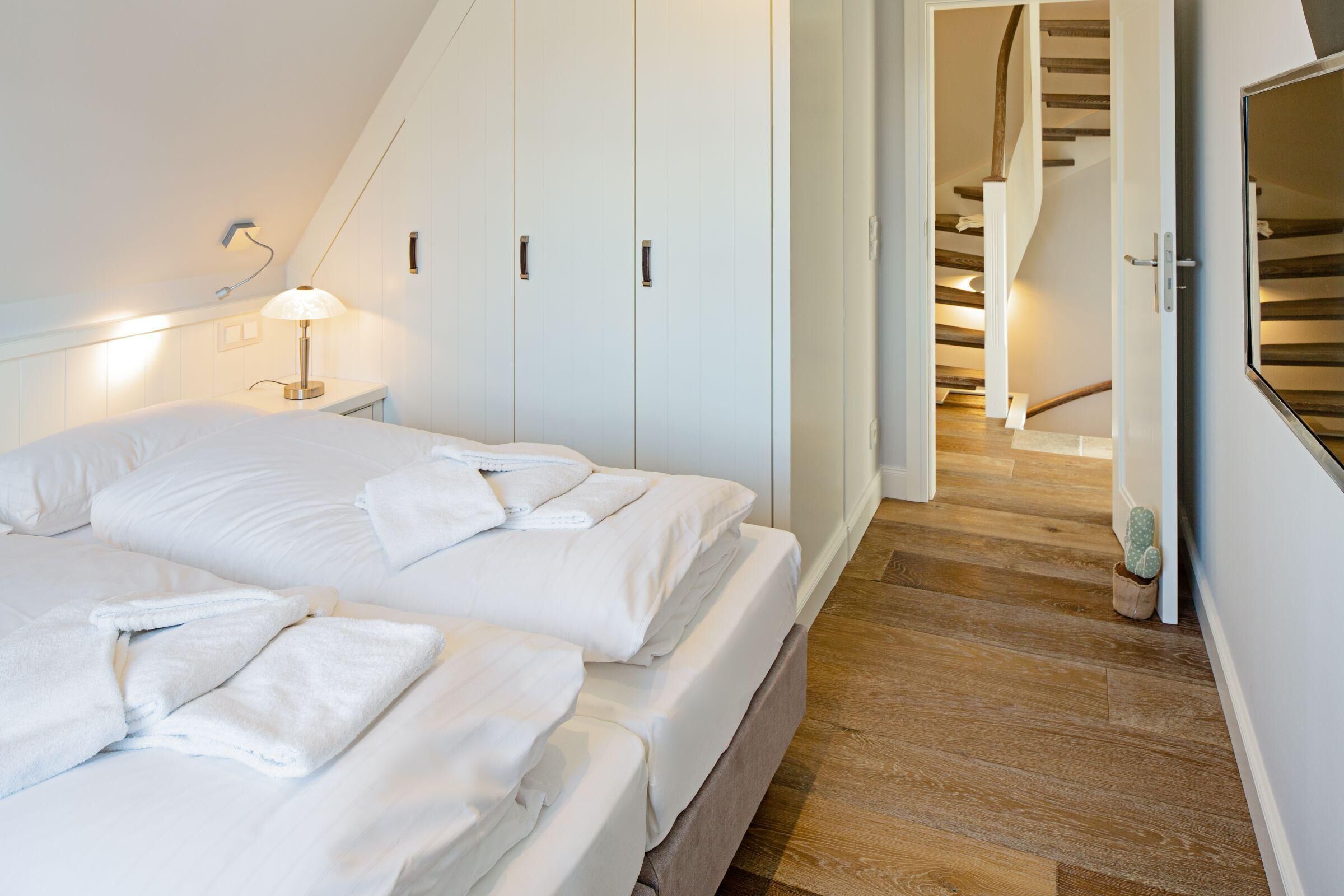 A bedroom with two beds, a nightstand with a lamp, white wardrobes, and a wooden staircase.