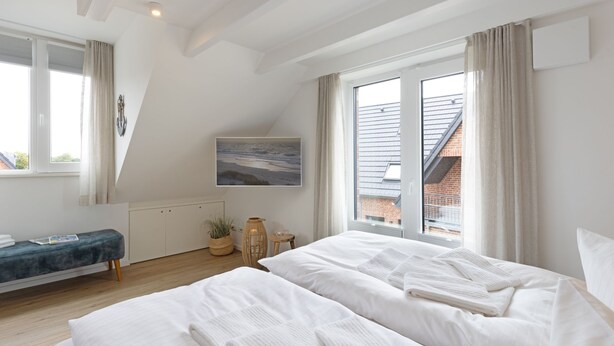 A bedroom with two beds, a bench, a painting, and a view of a brick building. - Herzlich Willkommen im Appartement Meerzeit (List)