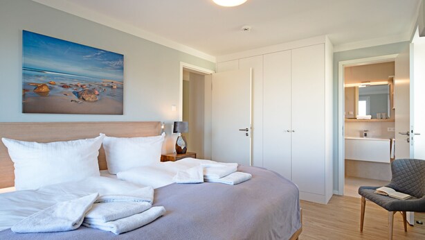 A neatly made bed with white linens and towels, a bedside lamp, and a painting of a beach scene on the wall. - Herzlich Willkommen im Appartement Priel (List)