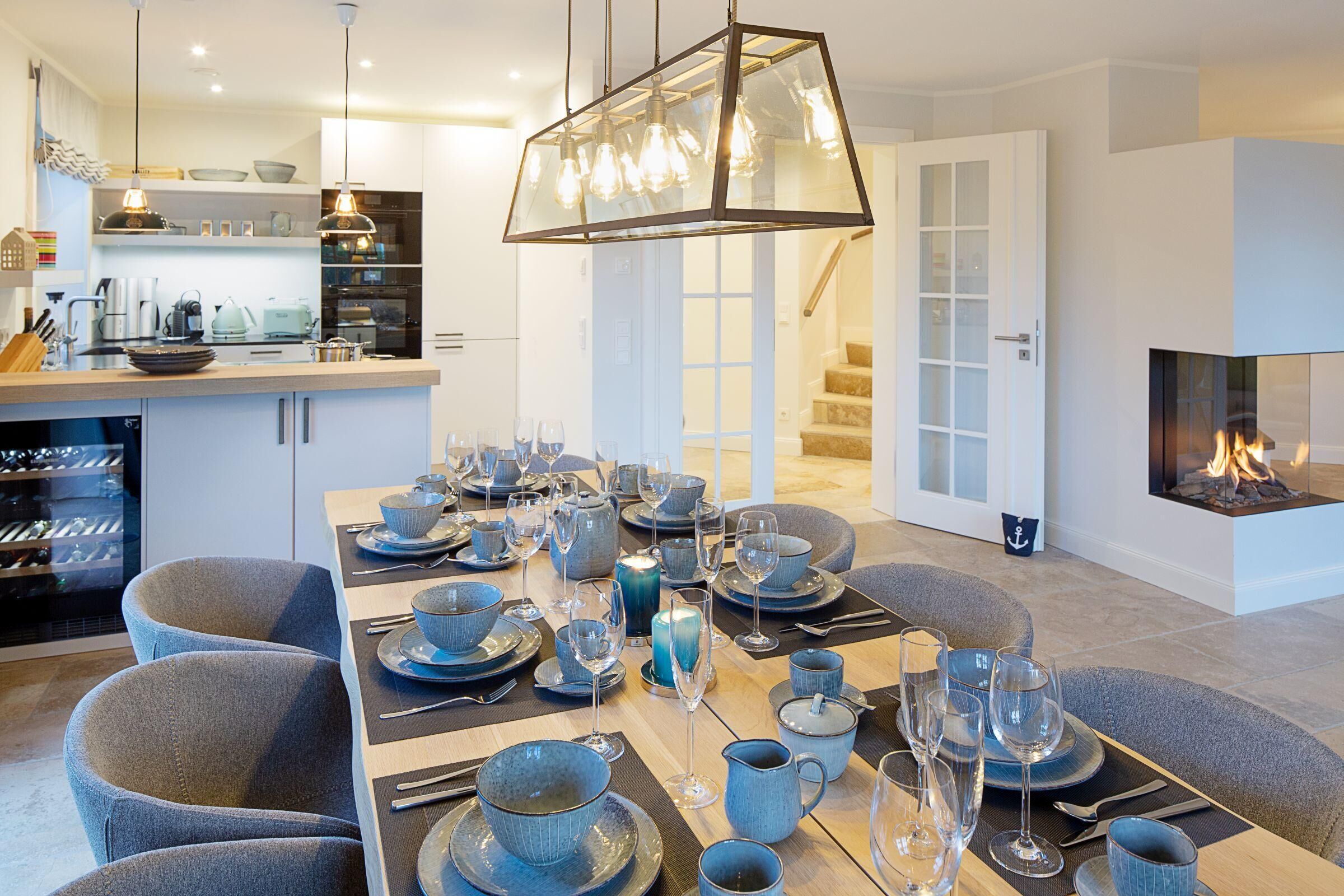 Un comedor moderno con una mesa larga dispuesta para una comida, una cocina al fondo y una chimenea.