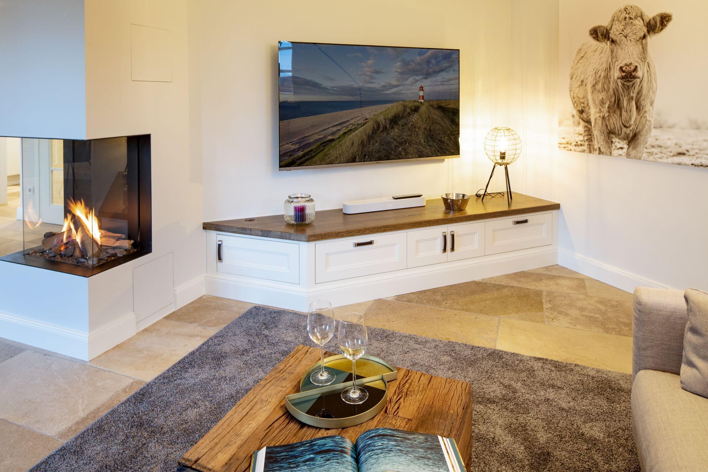 Una sala de estar moderna con chimenea, un televisor de pantalla plana montado en la pared, una mesa de centro de madera con un libro y vasos, y un cuadro grande de una vaca.
