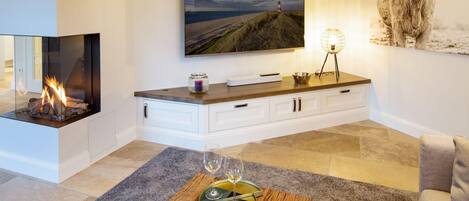 Una sala de estar moderna con chimenea, un televisor de pantalla plana montado en la pared, una mesa de centro de madera con un libro y vasos, y un cuadro grande de una vaca.