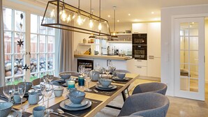 A modern dining area with a long table set for a meal, kitchen appliances in the background, and a view through large windows. - Herzlich Willkommen im Ferienhaus Trauminsel (List)