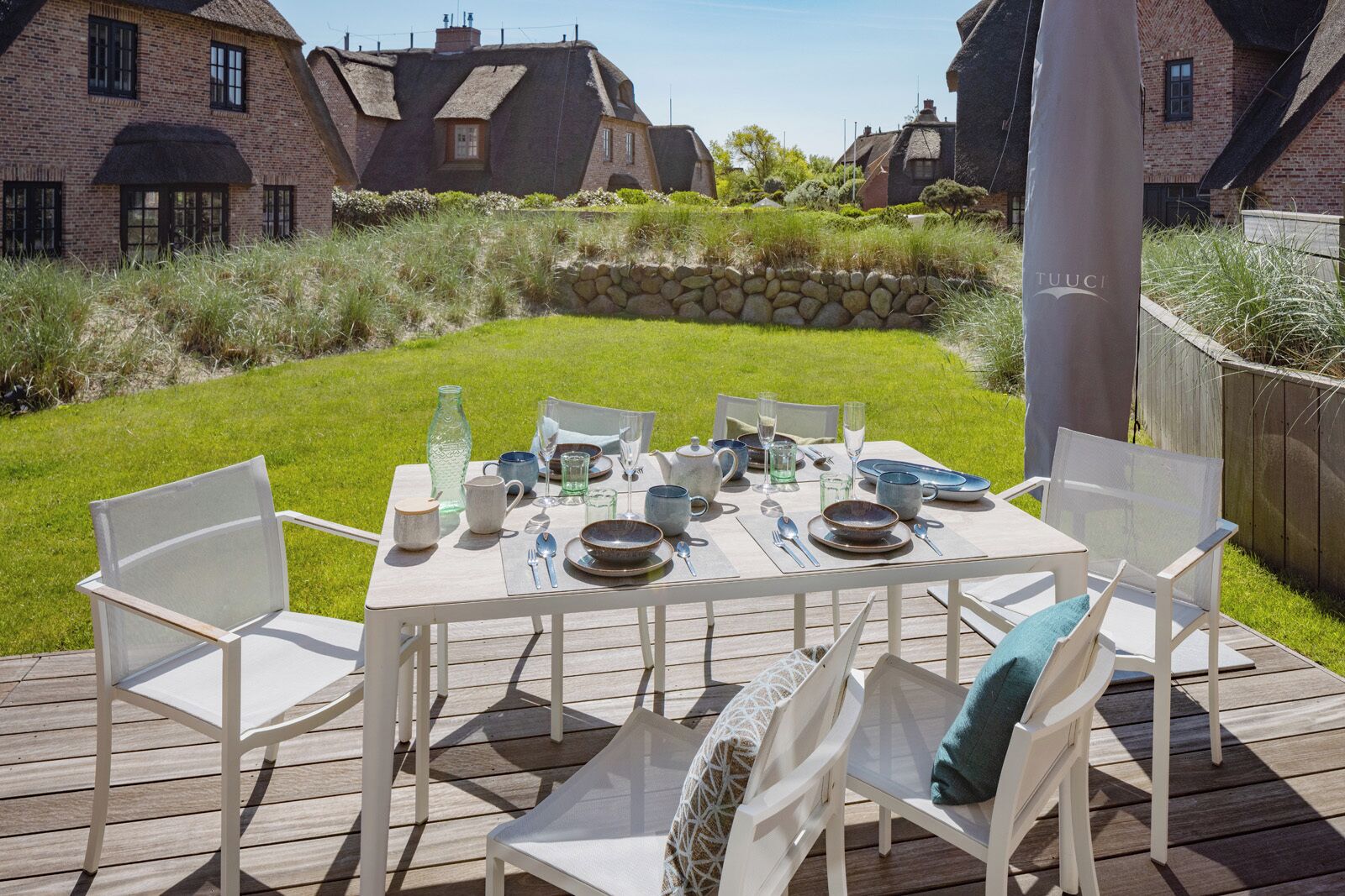 Um terraço com mesa posta para refeições, rodeado por cadeiras brancas, e um muro de pedra e casas ao fundo.