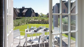 Een open terras met een eettafel, omringd door witte stoelen, met uitzicht op een tuin met een stenen muur en huizen op de achtergrond.