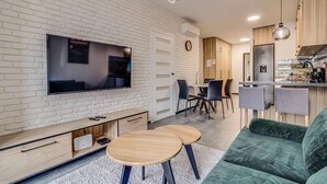 A modern living room with a flat-screen TV mounted on a white brick wall, a wooden entertainment unit, a green sofa, and a dining area with a round table and chairs. - RentPlanet - Apartament Wyszyńskiego (Wroclaw)