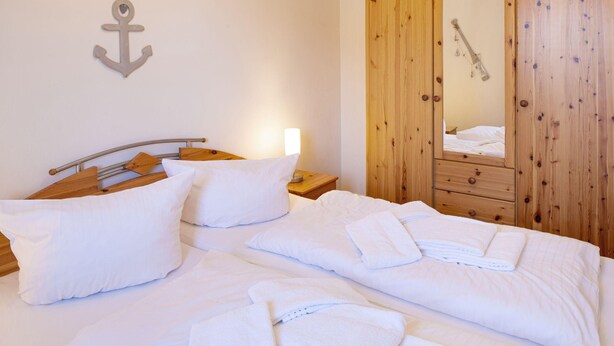Una cama bien hecha con sábanas y toallas blancas, cabecera de madera, mesita de noche con lámpara y armario de madera con espejo.