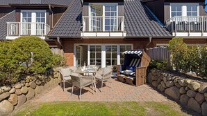 A two-story house with a patio area featuring a table and chairs, a covered seating area, and a stone wall. - Herzlich Willkommen im Ferienhaus Olivia (List)