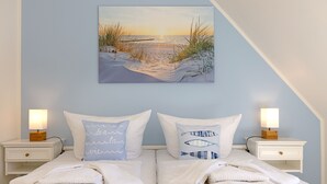 A bedroom with a bed, two nightstands, a painting of a beach scene, and two lamps. - Herzlich Willkommen im Ferienhaus Olivia (List)