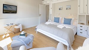 A bedroom with a large bed, a bedside table, a chair, a small table with a basket, and a wall-mounted TV. - Herzlich Willkommen im Appartement Heideblick (List)