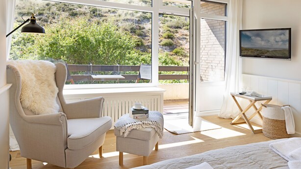 A bedroom with a large bed, a chair, a small table, a lamp, and a view of the outdoors. - Herzlich Willkommen im Appartement Heideblick (List)