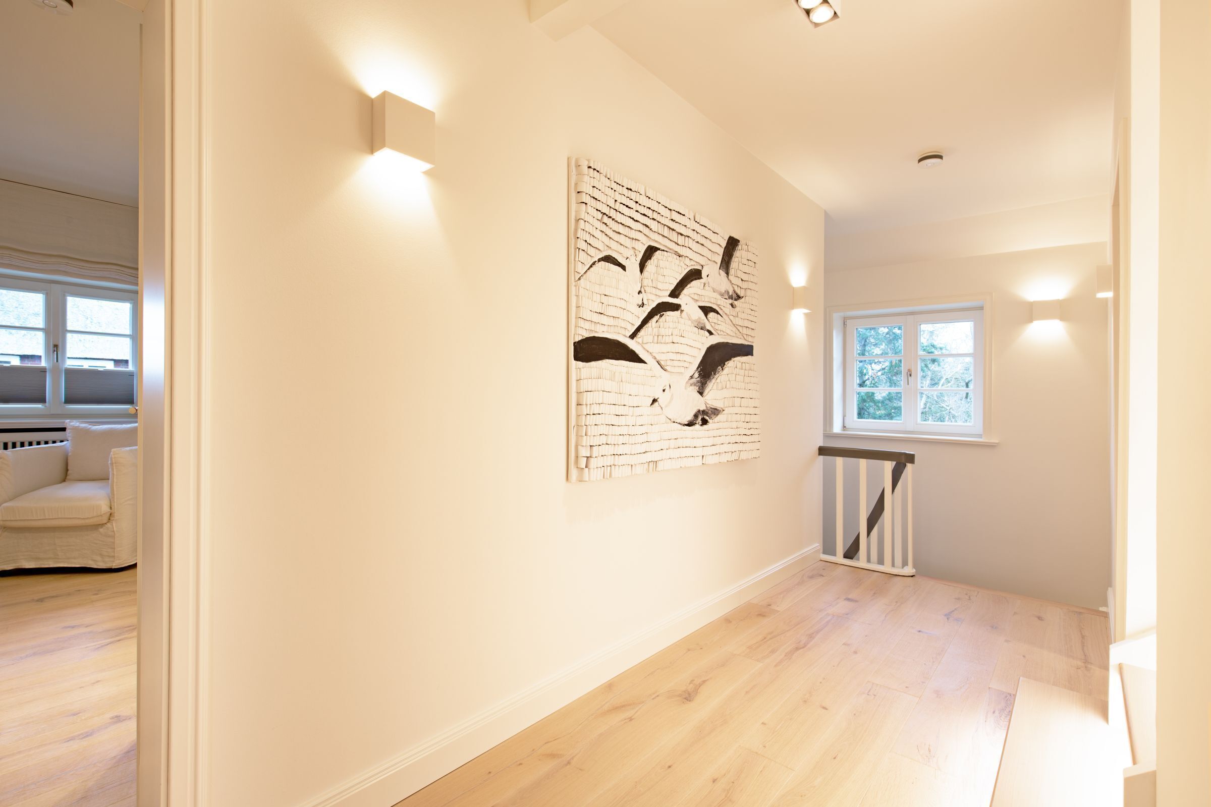 Un pasillo con piso de madera, pared blanca con una obra de arte texturizada, una ventana con persianas y una escalera con barandilla blanca.