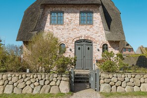 Una casa tradicional de ladrillo con tejado de paja, paredes de piedra y una entrada central con puerta y escalones.