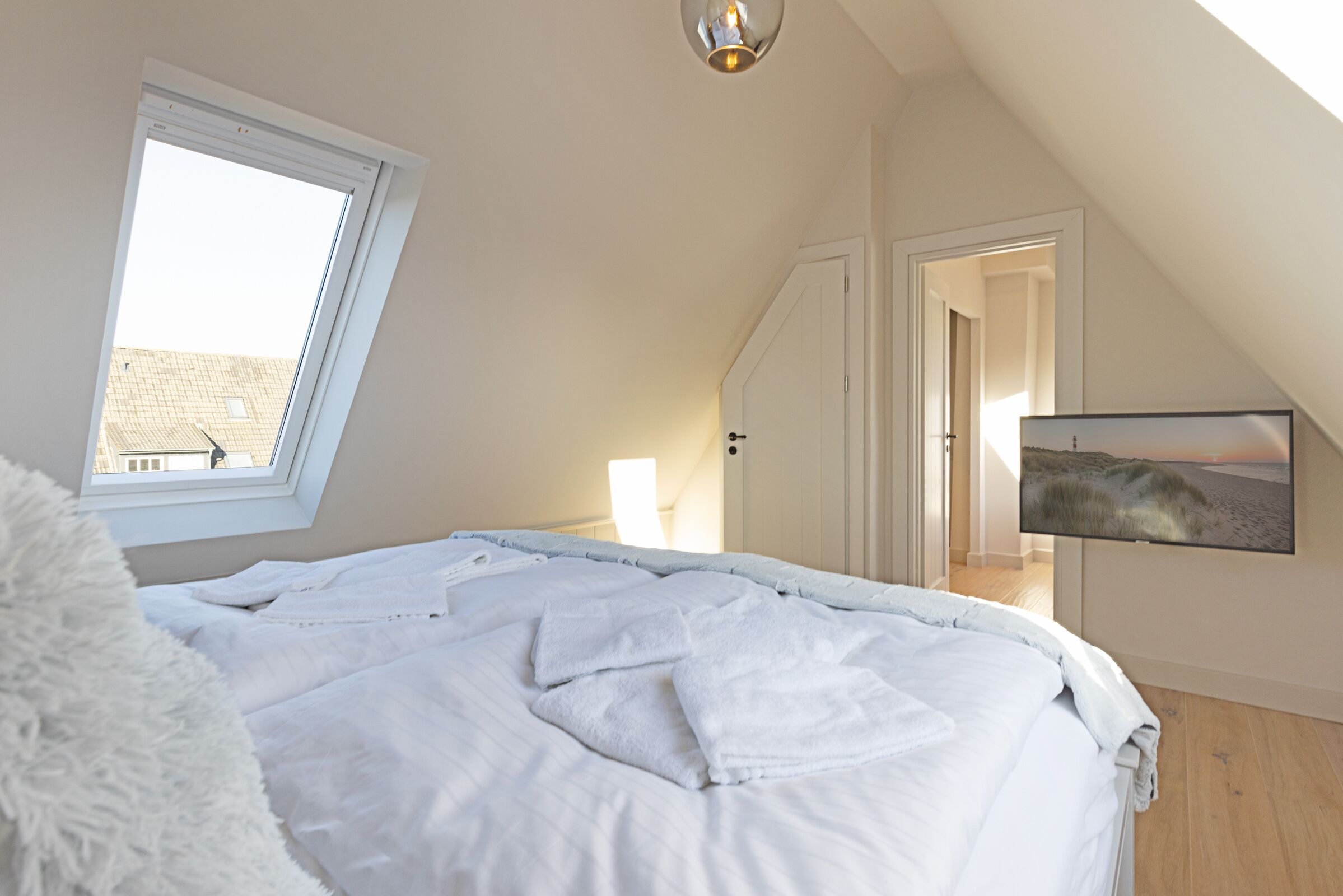 A bedroom with a skylight, a bed with white bedding, a TV on the wall, and a door leading to another room.
