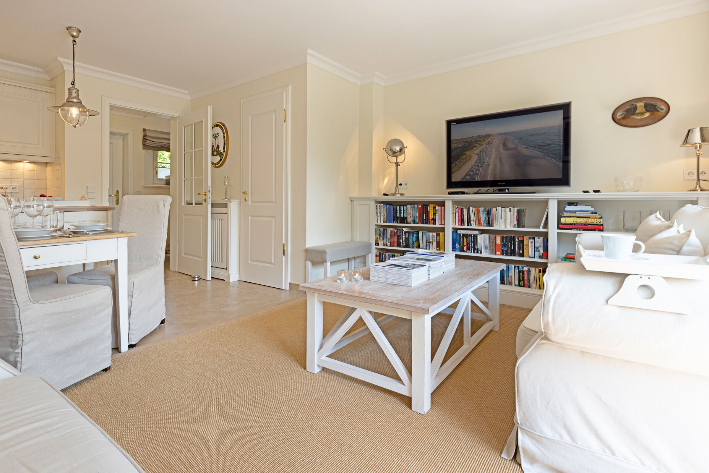 Una sala de estar con un sofá blanco, una mesa de centro, una estantería, un televisor y un comedor con mesa y sillas.