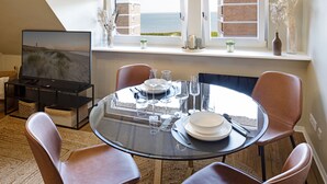 A dining area with a round glass table set for four, surrounded by leather chairs, with a television and a view of the sea visible through the windows.
