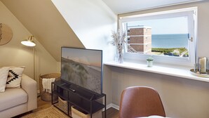 A modern living room with a TV, a dining table with plates, a window with a view of the sea, and a cozy seating area.