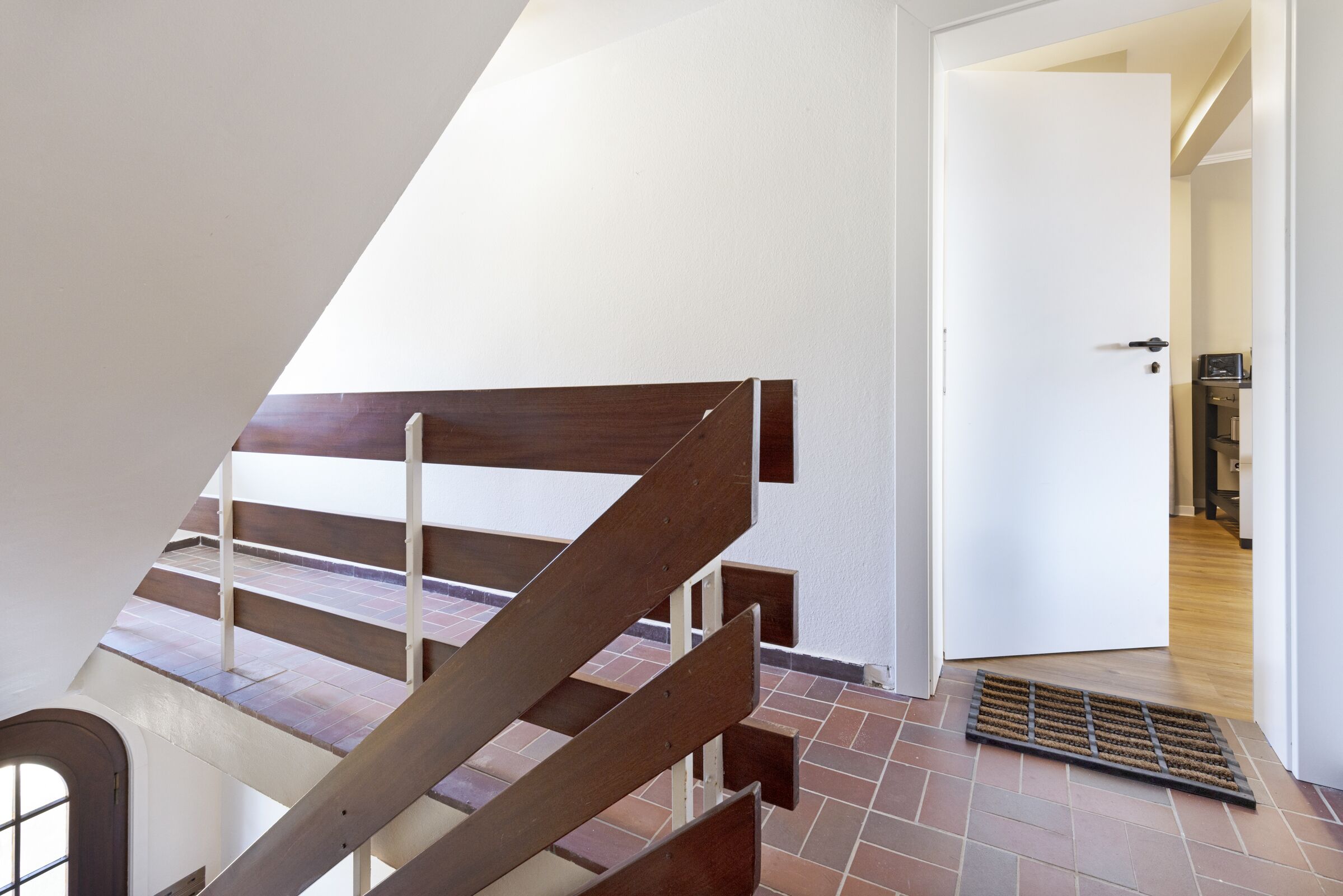 Un escalier avec des marches en bois et une rampe en métal, menant à une pièce dotée d’une porte blanche et d’un sol carrelé.