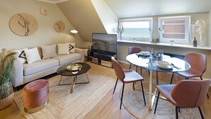 A modern living room with a sofa, coffee table, and dining area with chairs. There is a TV on a stand, a window with a view, and decorative wall art.