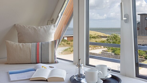 Une chambre dotée d’une grande fenêtre donnant sur la côte, un lit avec des oreillers, un plateau avec un livre, une tasse et un salier.