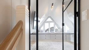 Un intérieur moderne avec un escalier en bois, un banc blanc agrémenté de coussins et une grande fenêtre donnant sur l’extérieur.