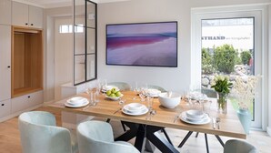 Un coin repas avec une table en bois dressée pour six personnes, entourée de chaises de couleur claire. Un grand tableau est accroché au mur au-dessus de la table, et une fenêtre offre une vue sur la verdure et un bâtiment.