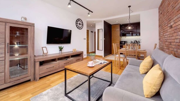 A modern living room with a grey sofa, wooden entertainment unit, and a flat-screen TV. - Industrial Apartment Fredry with Parking by Noclegi Renters (Kraków)