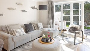 Una sala de estar moderna con un sofá gris, un reposapiés y una mesa de centro con jarrones. Se observan obras de arte de peces en la pared, un ventanal grande y una alfombra estampada.
