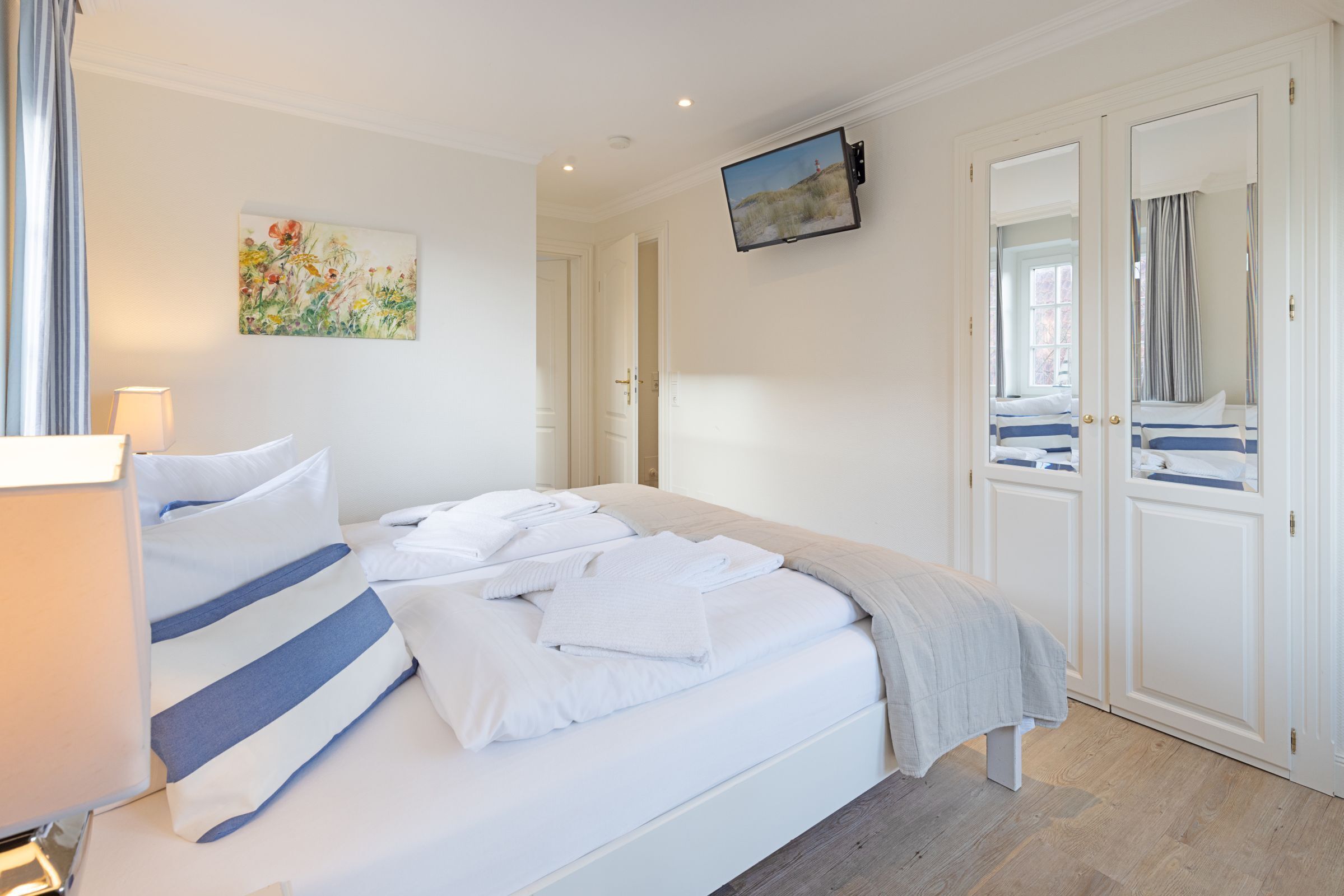 Un lit bien fait, orné d’oreillers à rayures blanches et bleues, un téléviseur à écran plat fixé au mur et un tableau représentant des fleurs accroché au mur.