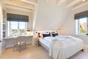 A bedroom with a sloped ceiling, a bed with white bedding, a desk with a chair, and two windows with curtains.