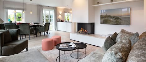 A modern living room with a fireplace, a dining area with a table and chairs, and a large window with curtains.