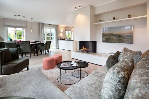 A modern living room with a fireplace, a dining area with a table and chairs, and a large window with curtains.