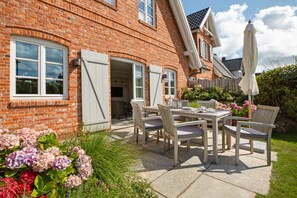 Ein Ziegelhaus mit einer Terrasse, ausgestattet mit Tisch und Stühlen, umgeben von üppigem Grün und Blumen.