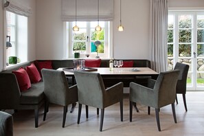 Un espace repas avec une longue table, plusieurs chaises et un banc. On y trouve des fenêtres avec des rideaux, une lampe et une vue sur la verdure à l’extérieur.