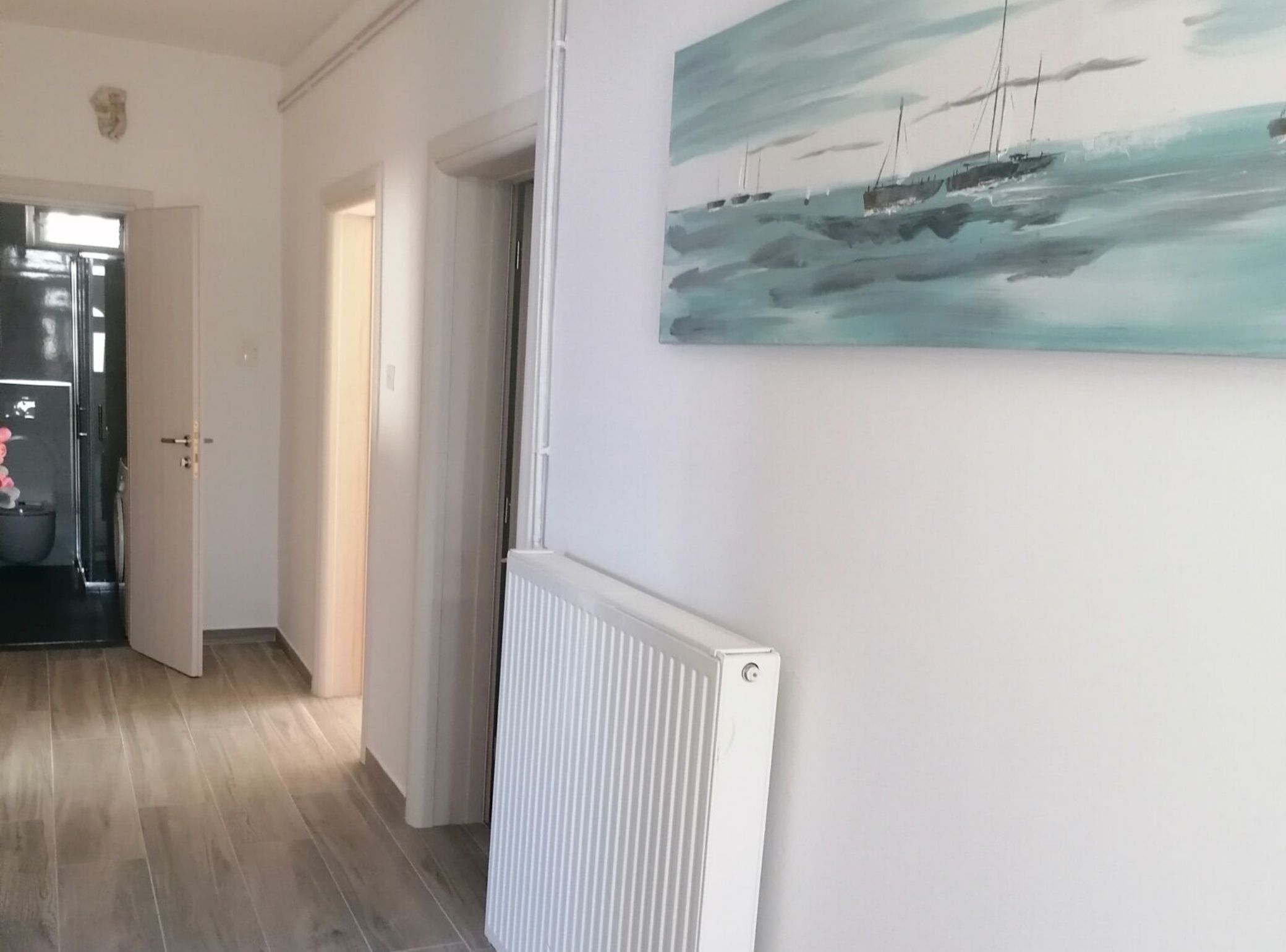 A hallway with a white radiator, a door leading to another room, and a painting of a seascape on the wall.