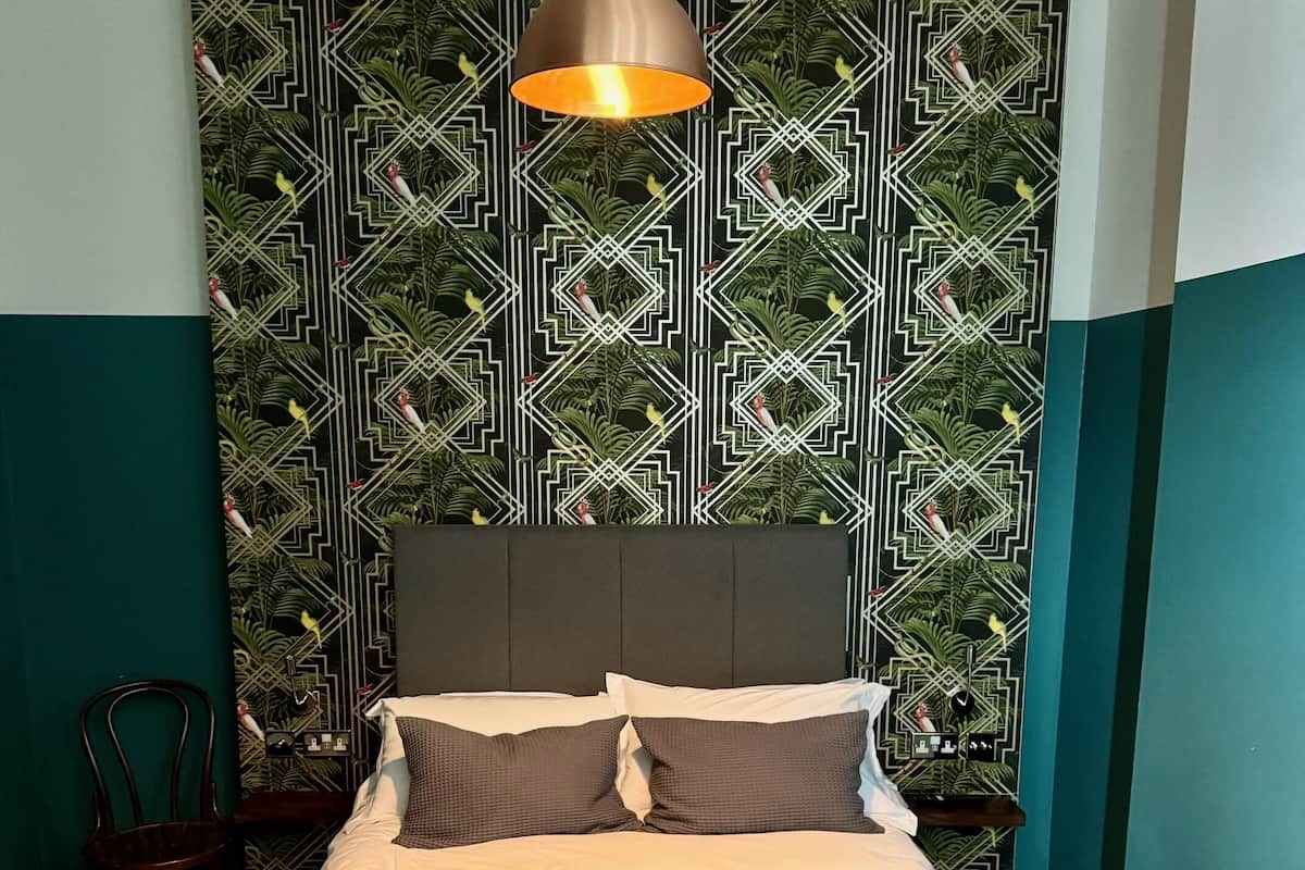 A bedroom with a patterned wallpaper, a bed with a headboard, a hanging light fixture, and a chair.