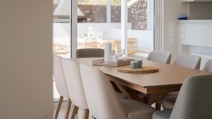 Uma sala de jantar com uma mesa de madeira, cadeiras bege e vista para o exterior através de janelas amplas.