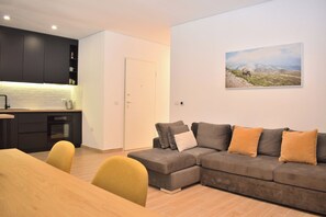 A modern living room with a grey sectional sofa, a wooden dining table, and a kitchen area with dark cabinets and white subway tiles.