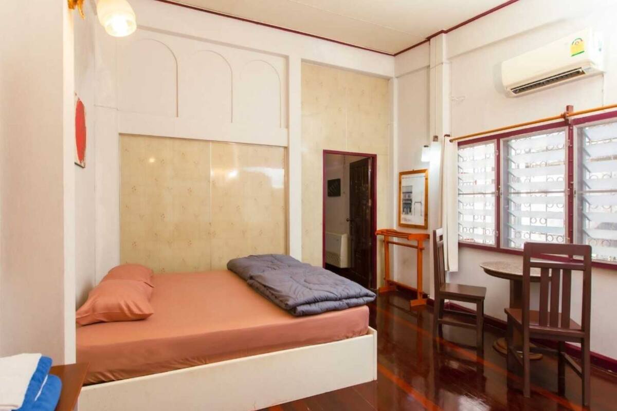 A room with a bed, a desk, and a chair. There is a window with blinds, an air conditioning unit, and a door leading to another room.