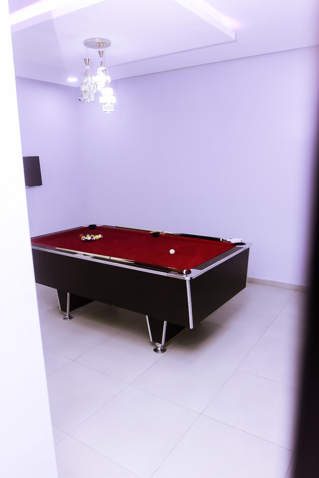 A pool table with a red felt surface, a chandelier, and a white tiled floor in a room with white walls.