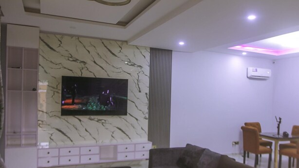 A modern living room with a marble wall, a flat-screen TV, a sofa, and a dining area with chairs. - Stunning 3-bed House in Lagos (Lagos)