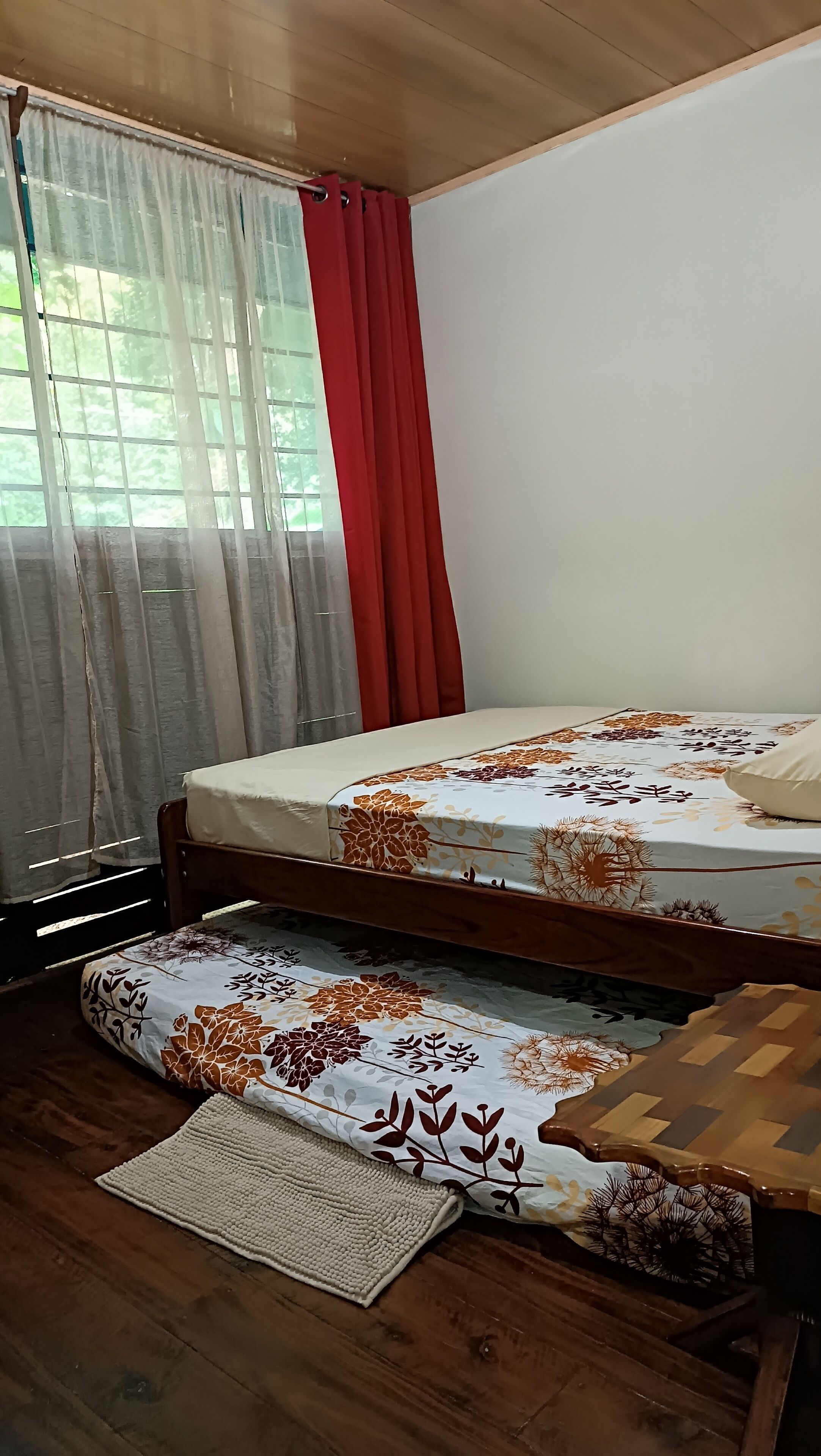 A room with a bed, a wooden floor, a window with curtains, and a view of greenery outside.